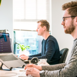 Two men collaborating at a desk, focused on their computers, surrounded by developer resources and tools.