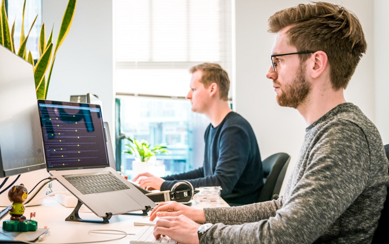 Two men collaborating at a desk, focused on their computers, surrounded by developer resources and tools.